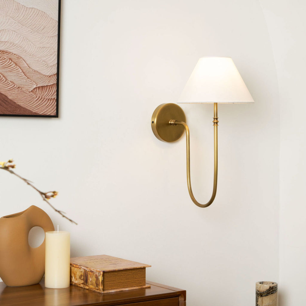 Brass and white Clérachic wall light on a pale wall, with a looping arm, a framed print at the left and a candle on a cabinet below.