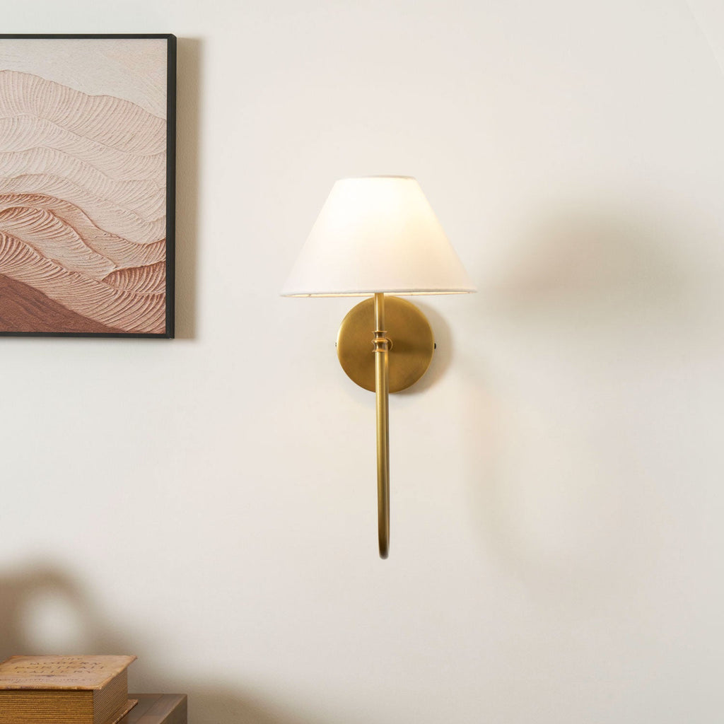 A brass Clérachic wall light with a white shade and a straight lower arm, with a framed print at the left and books on a cabinet below.