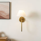 A brass Clérachic wall light with a white shade and a straight lower arm, with a framed print at the left and books on a cabinet below.