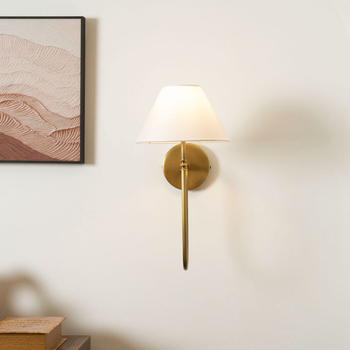 A brass Clérachic wall light with a white shade and a straight lower arm, with a framed print at the left and books on a cabinet below.