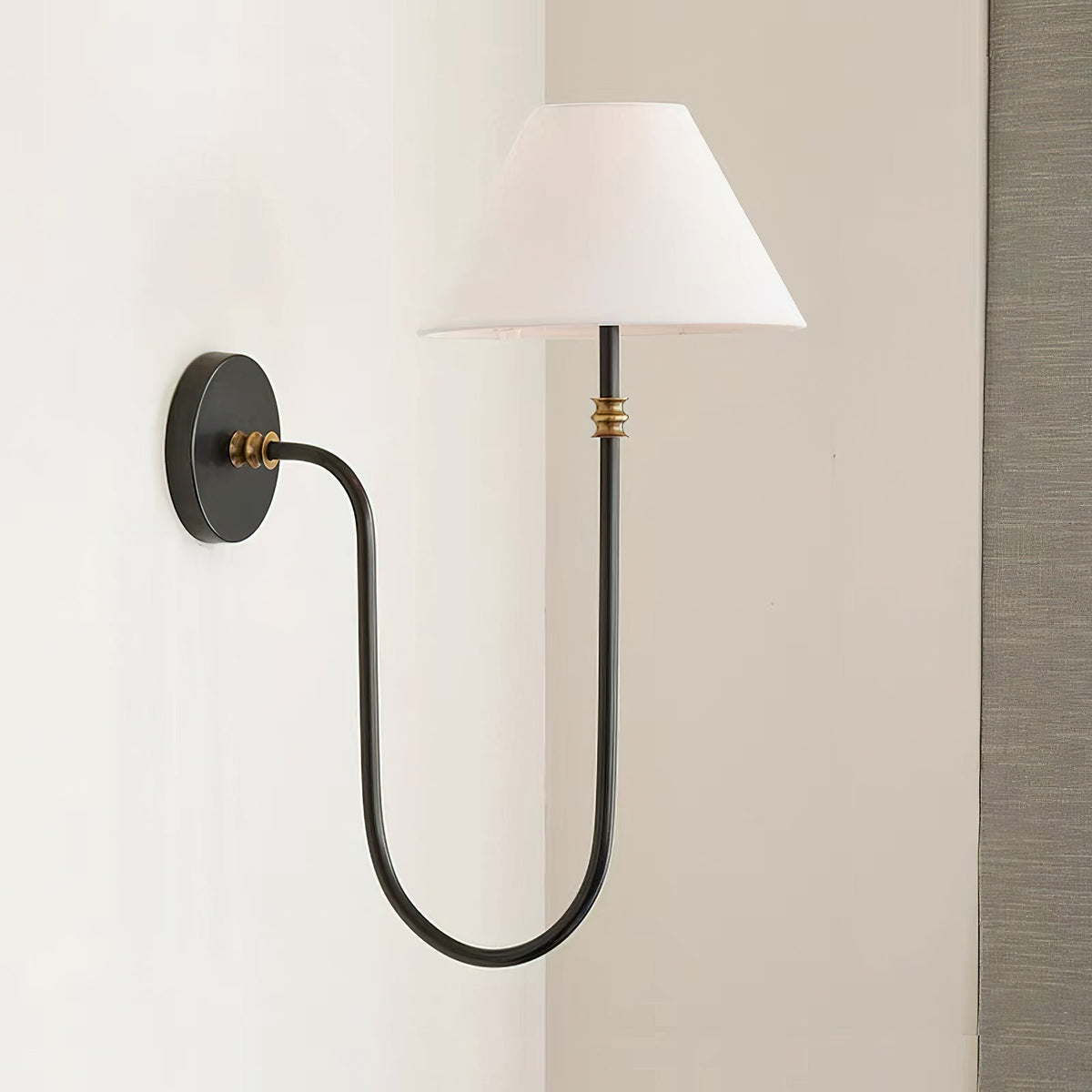 Side view of a black Clérachic wall light on a pale wall, with the U-shaped arm, small brass collar and white shade near a grey curtain.