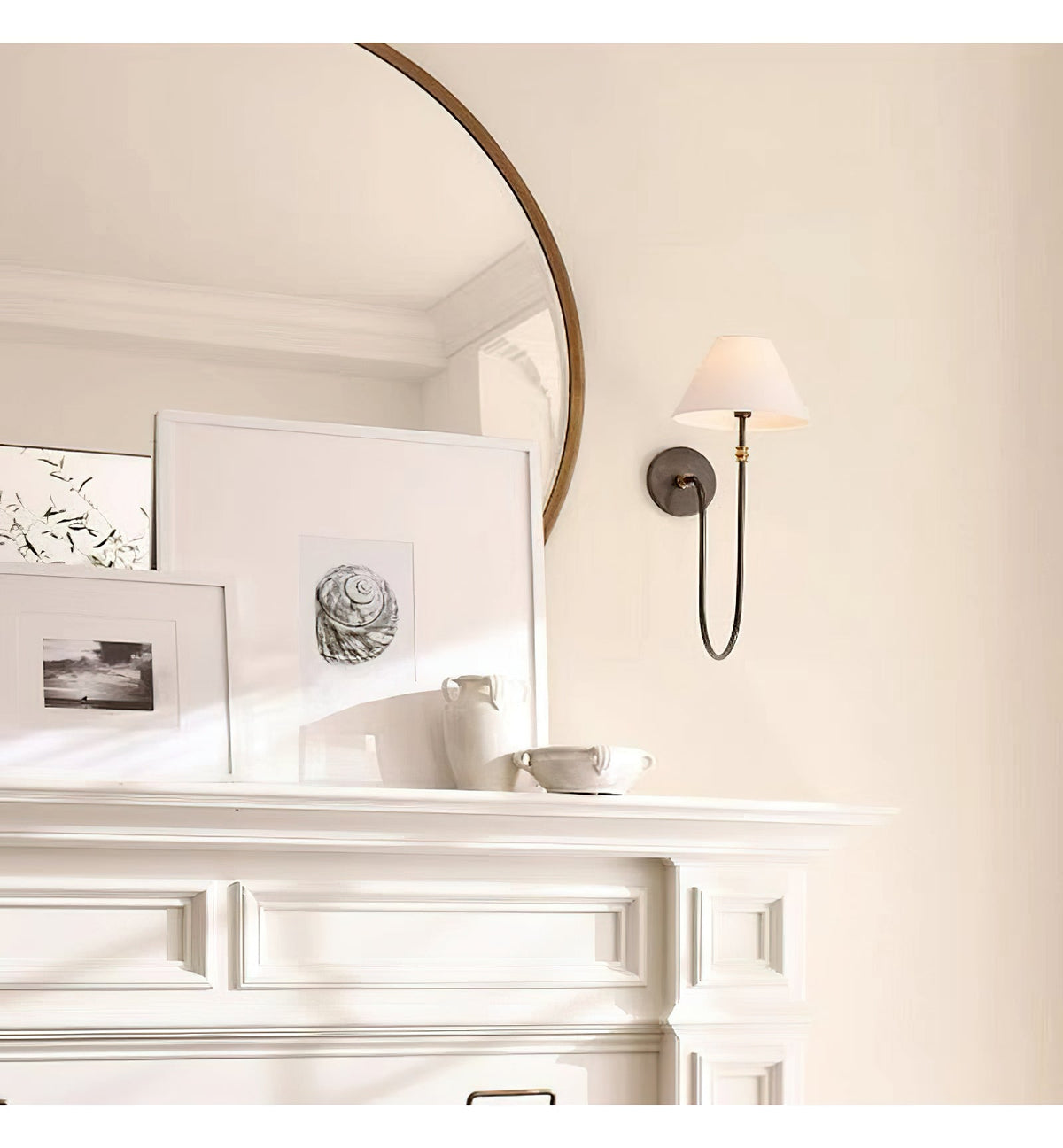 Distant view of a black Clérachic wall light with a white shade on a cream wall, beside a curved mirror above a white mantel.