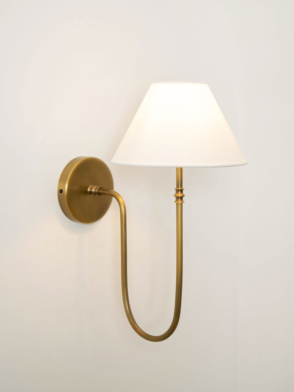 Brass Clérachic wall light with a white fabric shade lit warm, on a pale wall, with a round plate and a curved arm.