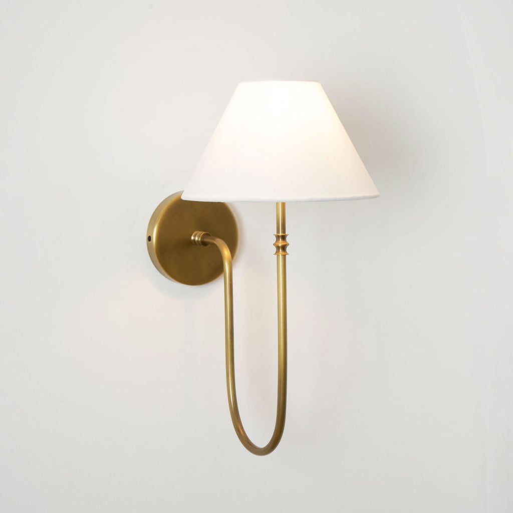 Aged brass Clérachic wall light with a white tapered shade, seen at an angle on a cream wall, with a long curved stem.