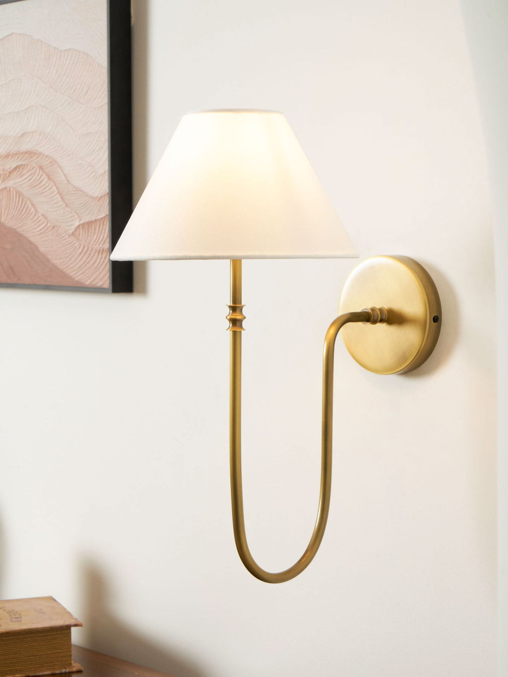 Brass Clérachic wall light on the right of a pale wall with a pink abstract print at the upper left and books below.
