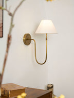 A brass Clérachic wall light with a white shade beside blossom branches, above a wooden cabinet with an old book and a small vase.