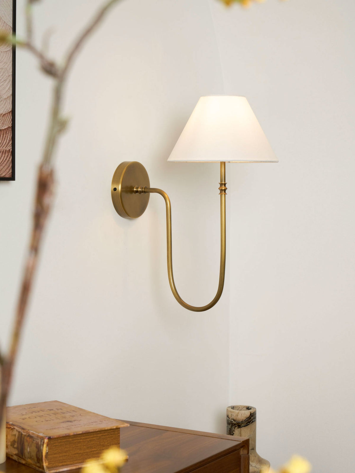 A brass Clérachic wall light with a white shade beside blossom branches, above a wooden cabinet with an old book and a small vase.