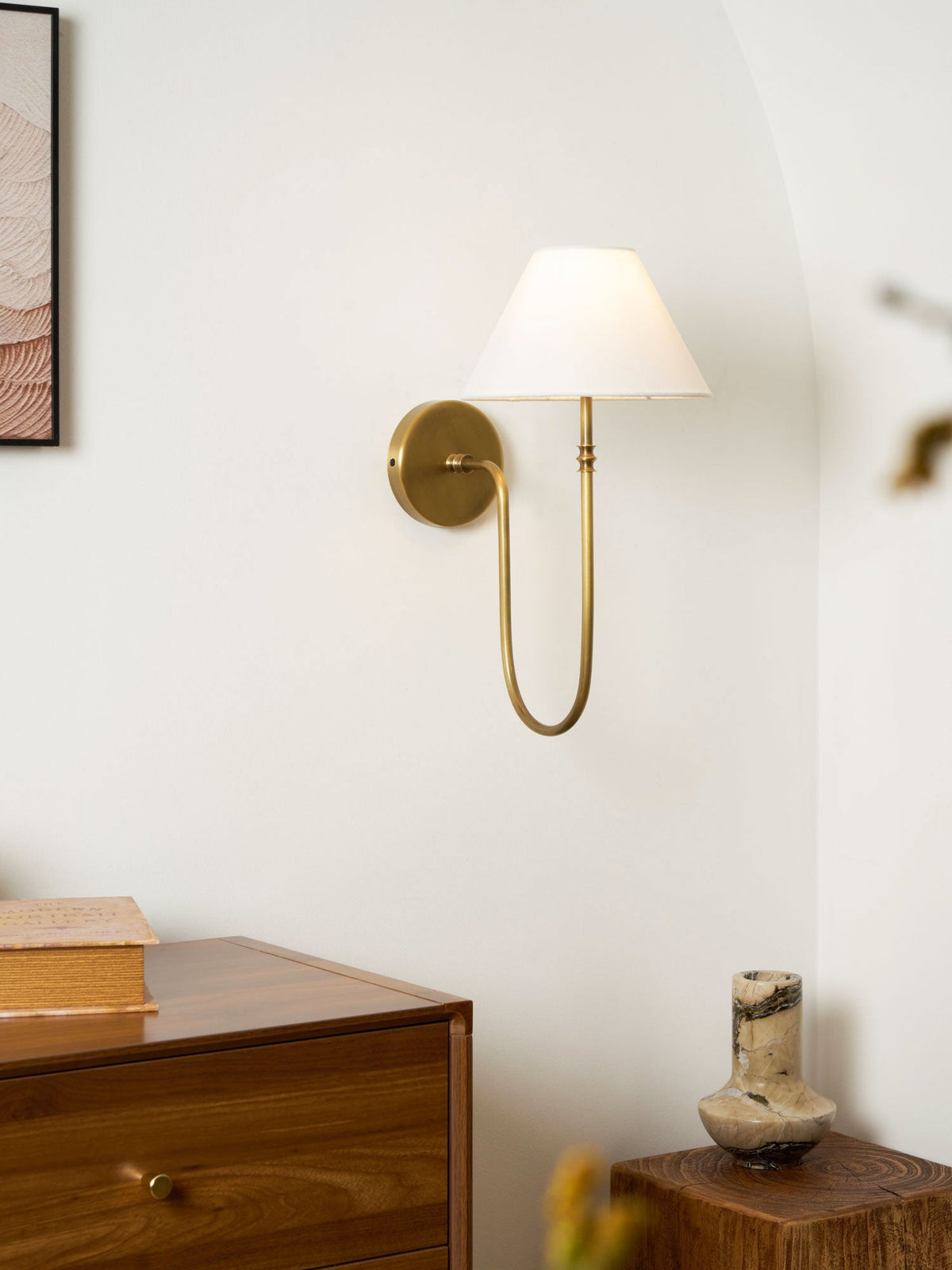 Smaller view of a brass Clérachic wall light with a white shade above a walnut chest of drawers and a stone vase.