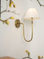 Brass Clérachic wall light with a lit white shade on a pale wall, framed by blossom branches with yellow buds.