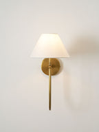 A brass Clérachic wall light with a straight lower arm and a white shade lit softly on a plain pale wall.