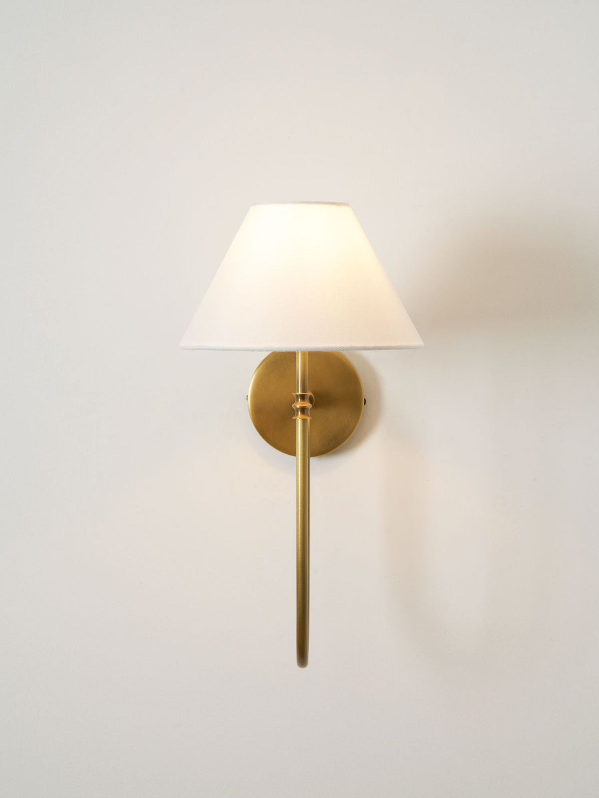 A brass Clérachic wall light with a straight lower arm and a white shade lit softly on a plain pale wall.