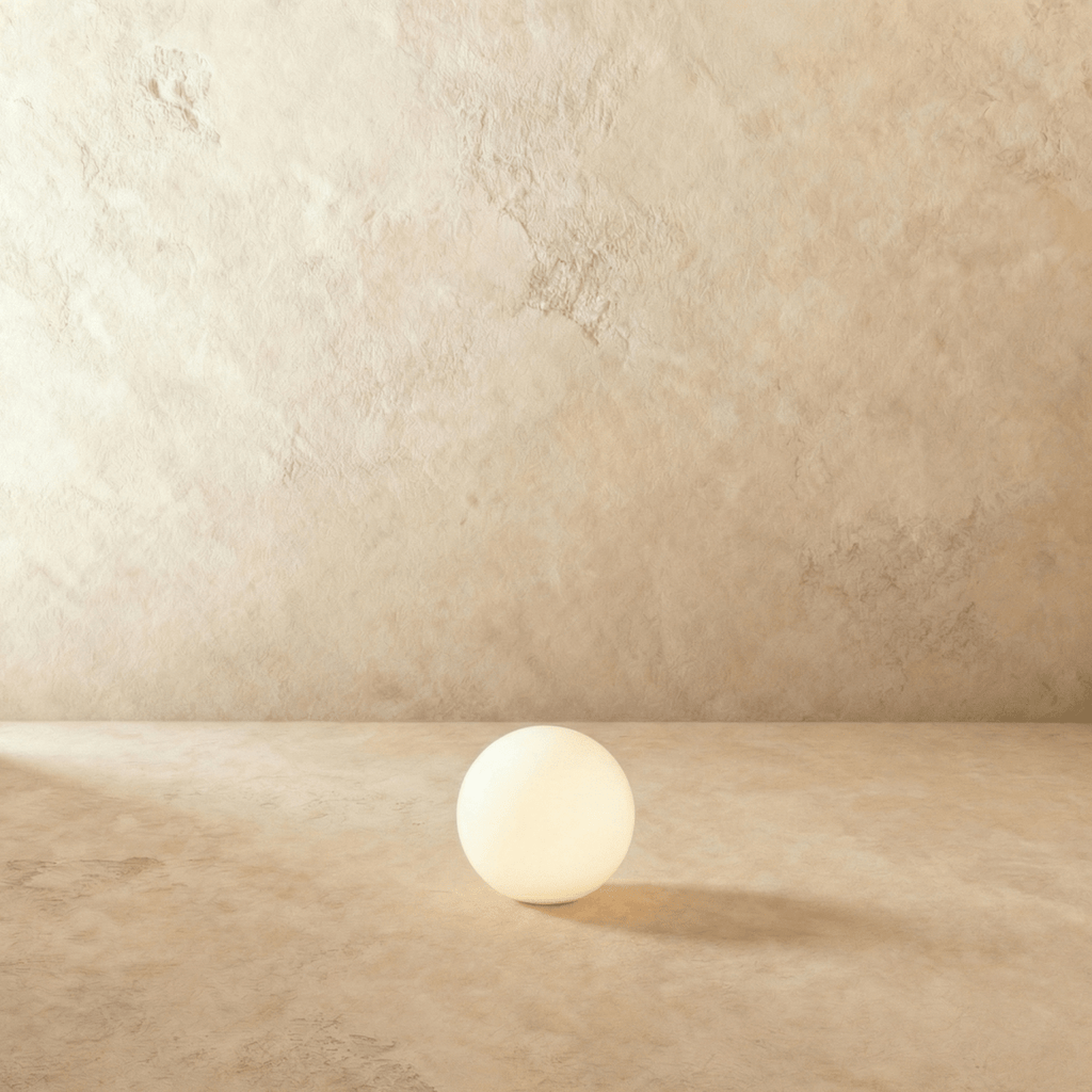 A Clérachic glowing sphere lamp glows softly on a plaster floor, its warm light pooling beneath the globe.