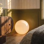 Close view of the lit Clérachic sphere floor lamp glowing on wood flooring beside a dresser and checked bedding.