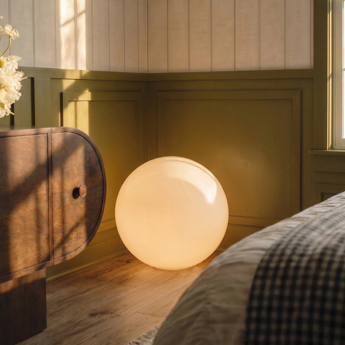 Close view of the lit Clérachic sphere floor lamp glowing on wood flooring beside a dresser and checked bedding.