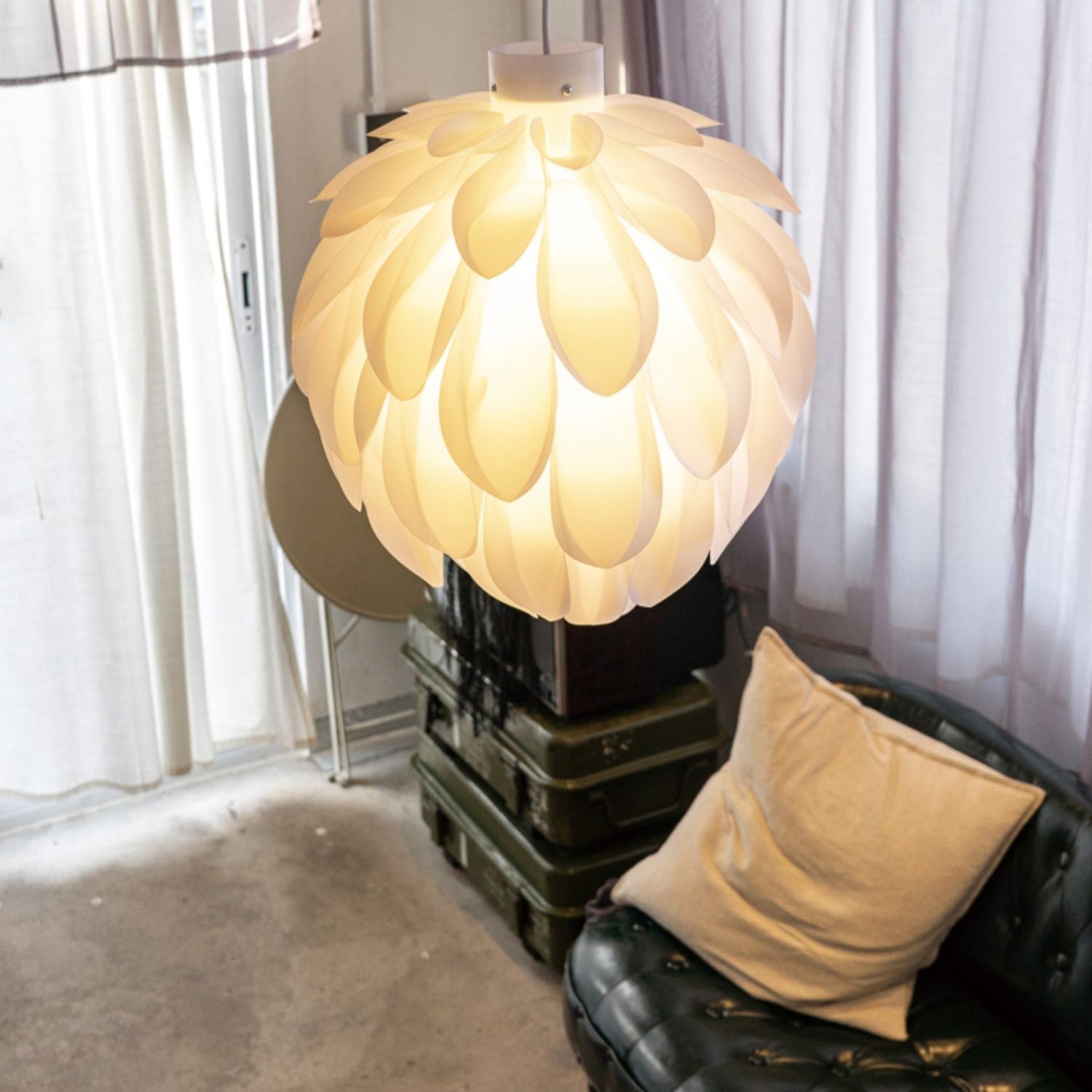 Clérachic White Flower Pendant Light glows beside a black tufted sofa and stacked olive storage trunks.