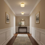Warm light from two Clérachic flush-mount lights along a beige hallway with white wainscoting, a console table and framed prints at the end.
