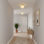 A Clérachic flush-mount ceiling light in a pale grey hallway, with a light oak console, a potted palm and two framed abstract prints.