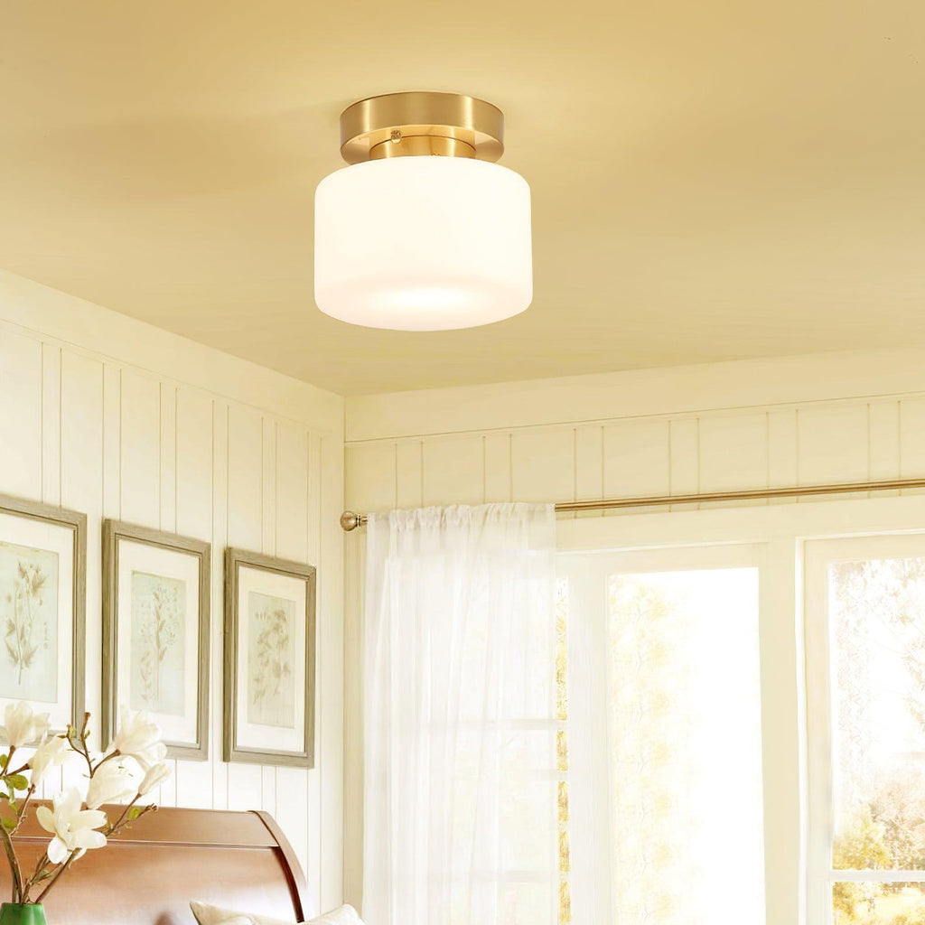 A Clérachic flush-mount light with a brass canopy above a bedroom corner, with framed prints, a wooden headboard and sheer curtains beyond.