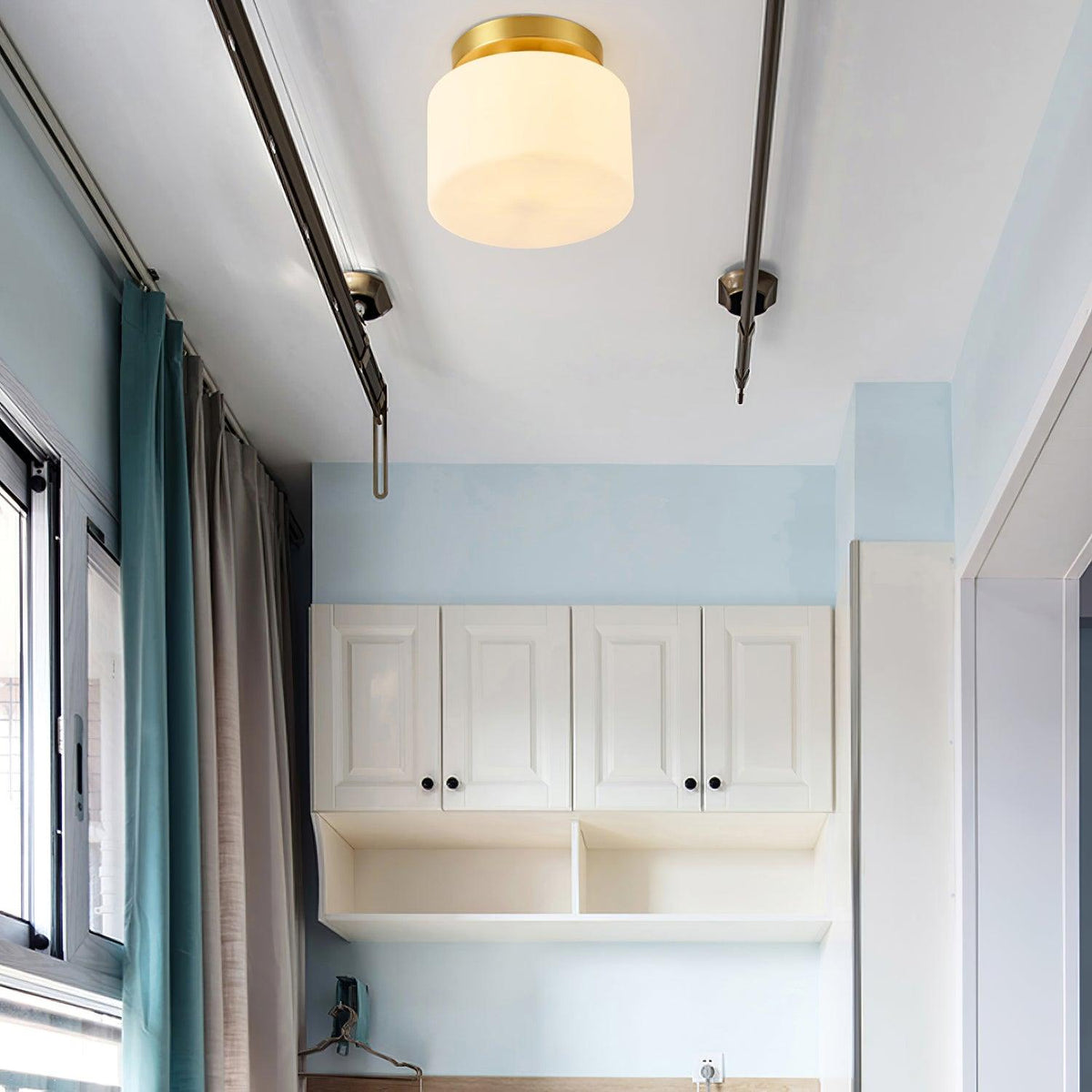 A Clérachic flush-mount light on a pale ceiling next to dark curtain rails, with a blue wall, curtains and white cabinets in the room.