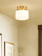 A Clérachic brass and white glass flush-mount light in a corner room with grasscloth-style walls, a dark door frame and a framed print.