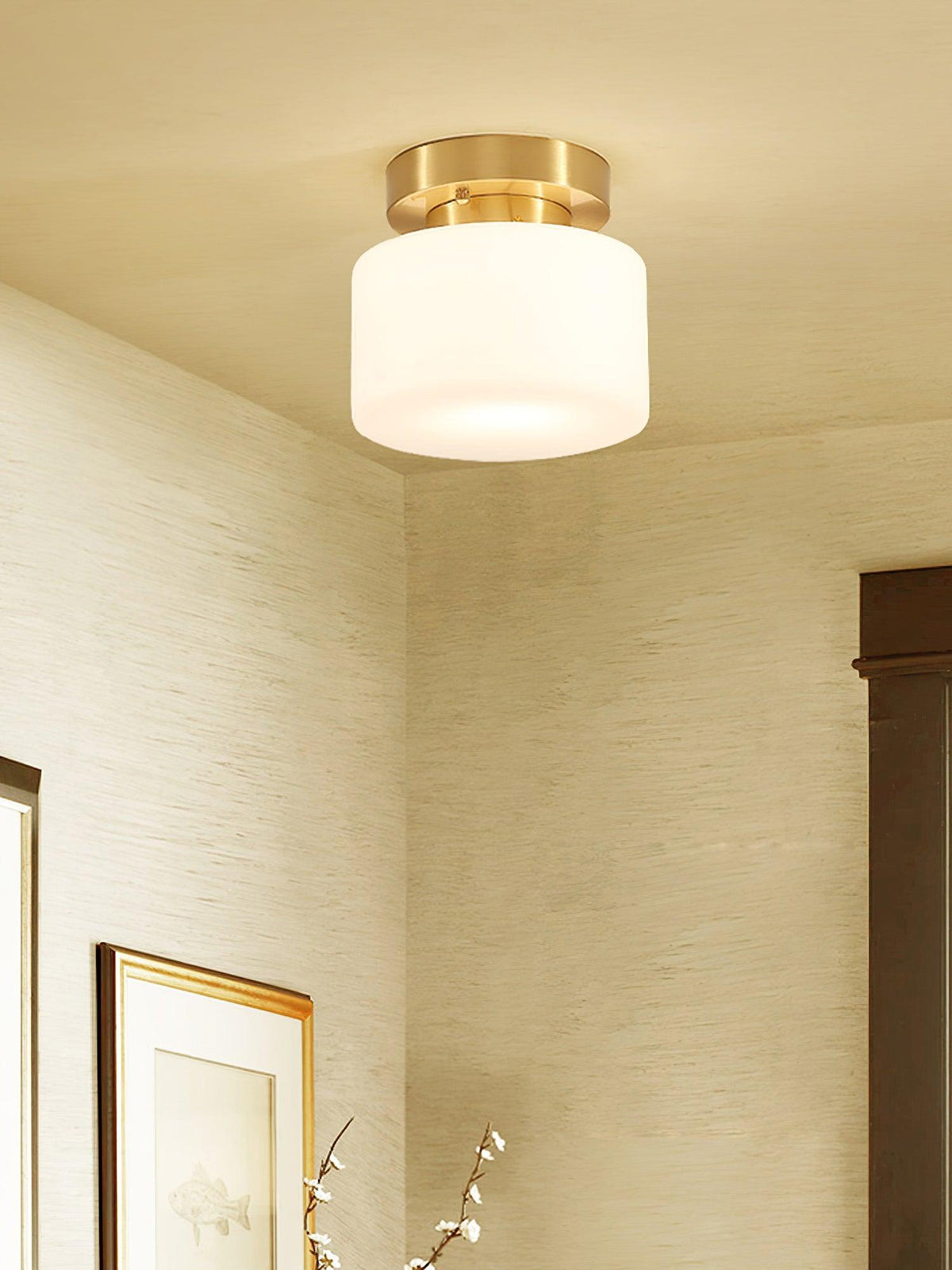A Clérachic brass and white glass flush-mount light in a corner room with grasscloth-style walls, a dark door frame and a framed print.