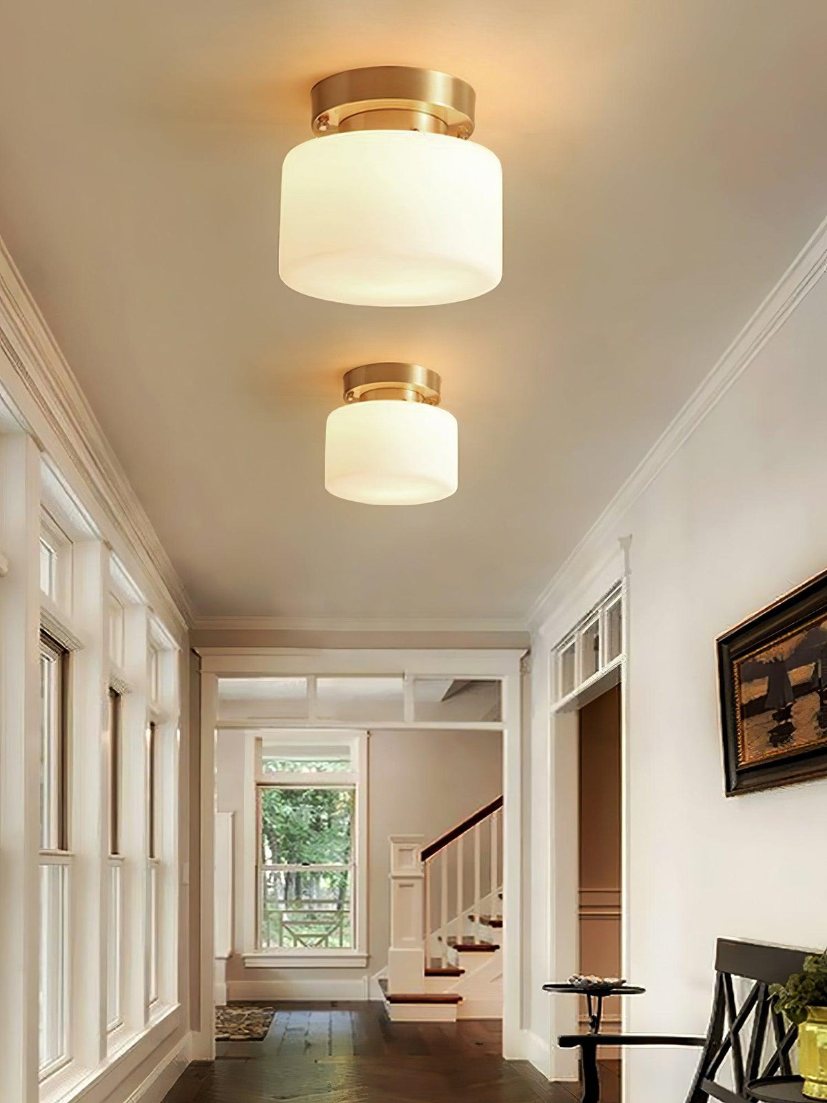 Two Clérachic flush-mount lights over a white hallway with tall windows, a view of green trees and a staircase in the distance.