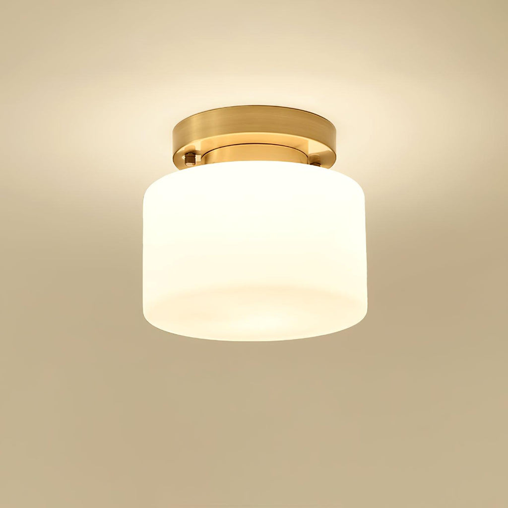 A Clérachic white glass drum flush-mount light lit warmly, with a soft glow on a golden-beige ceiling around the brass canopy.