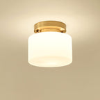 A Clérachic white glass drum flush-mount light lit warmly, with a soft glow on a golden-beige ceiling around the brass canopy.