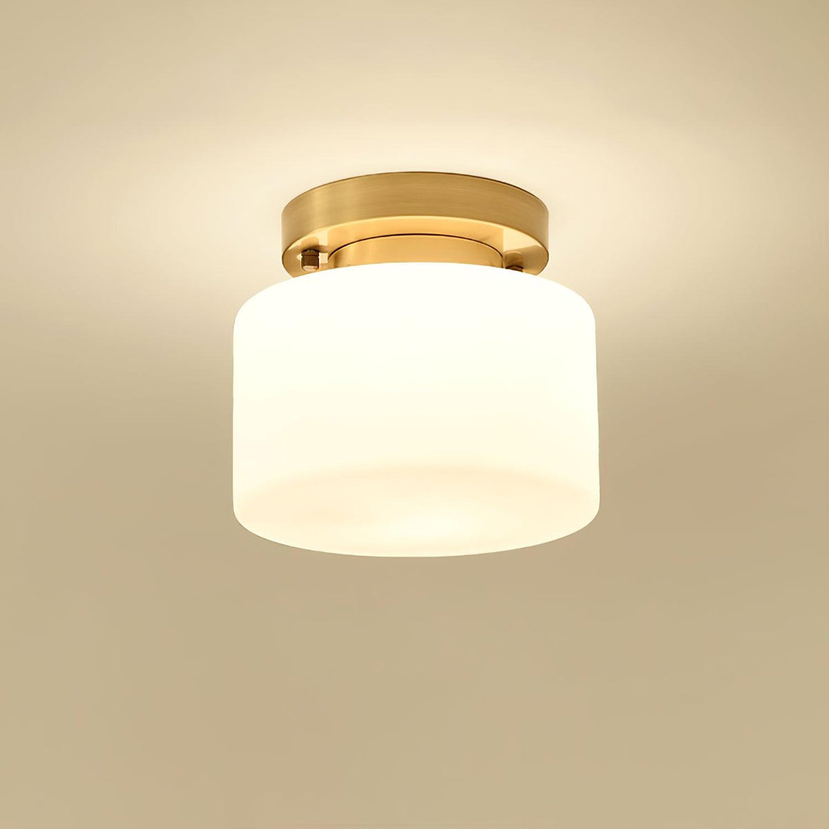 A Clérachic white glass drum flush-mount light lit warmly, with a soft glow on a golden-beige ceiling around the brass canopy.