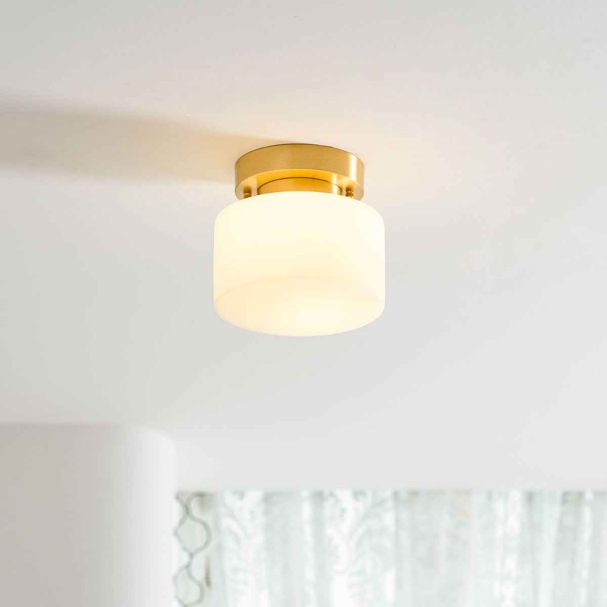 A Clérachic flush-mount light glowing softly on a white ceiling in a bright room, with a sheer curtain and pale walls behind.