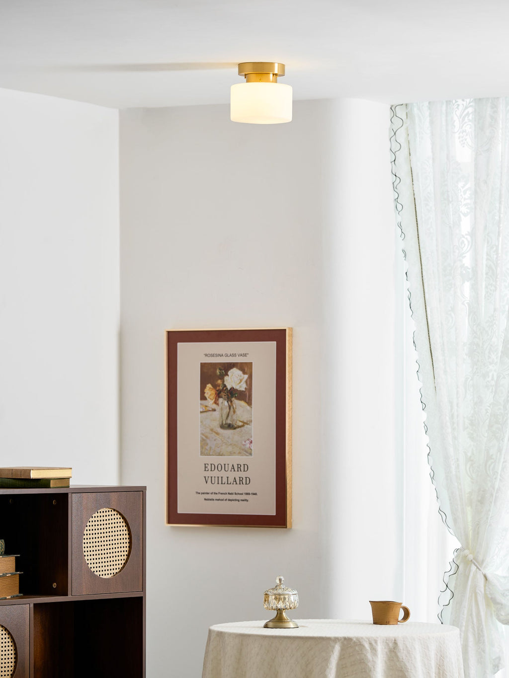 A Clérachic flush-mount light seen from afar on a white ceiling above a round table, with a framed print and a lace curtain.