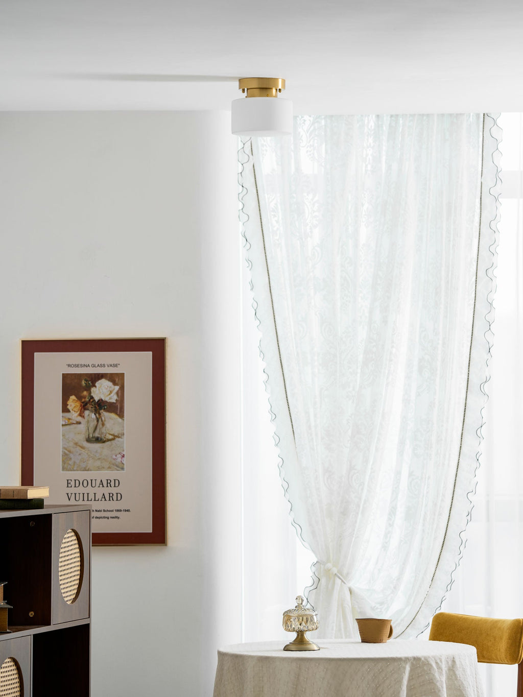 A Clérachic flush-mount light on a white ceiling with a sheer curtain, a table with a cup and a wooden shelf unit in a bright corner.