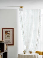 A Clérachic flush-mount light on a white ceiling with a sheer curtain, a table with a cup and a wooden shelf unit in a bright corner.