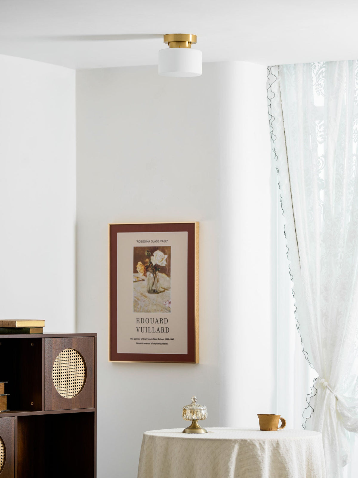 Wider view of a bright room with a Clérachic flush-mount light on the ceiling, a framed print, a rattan cabinet and a table with a lace cloth.