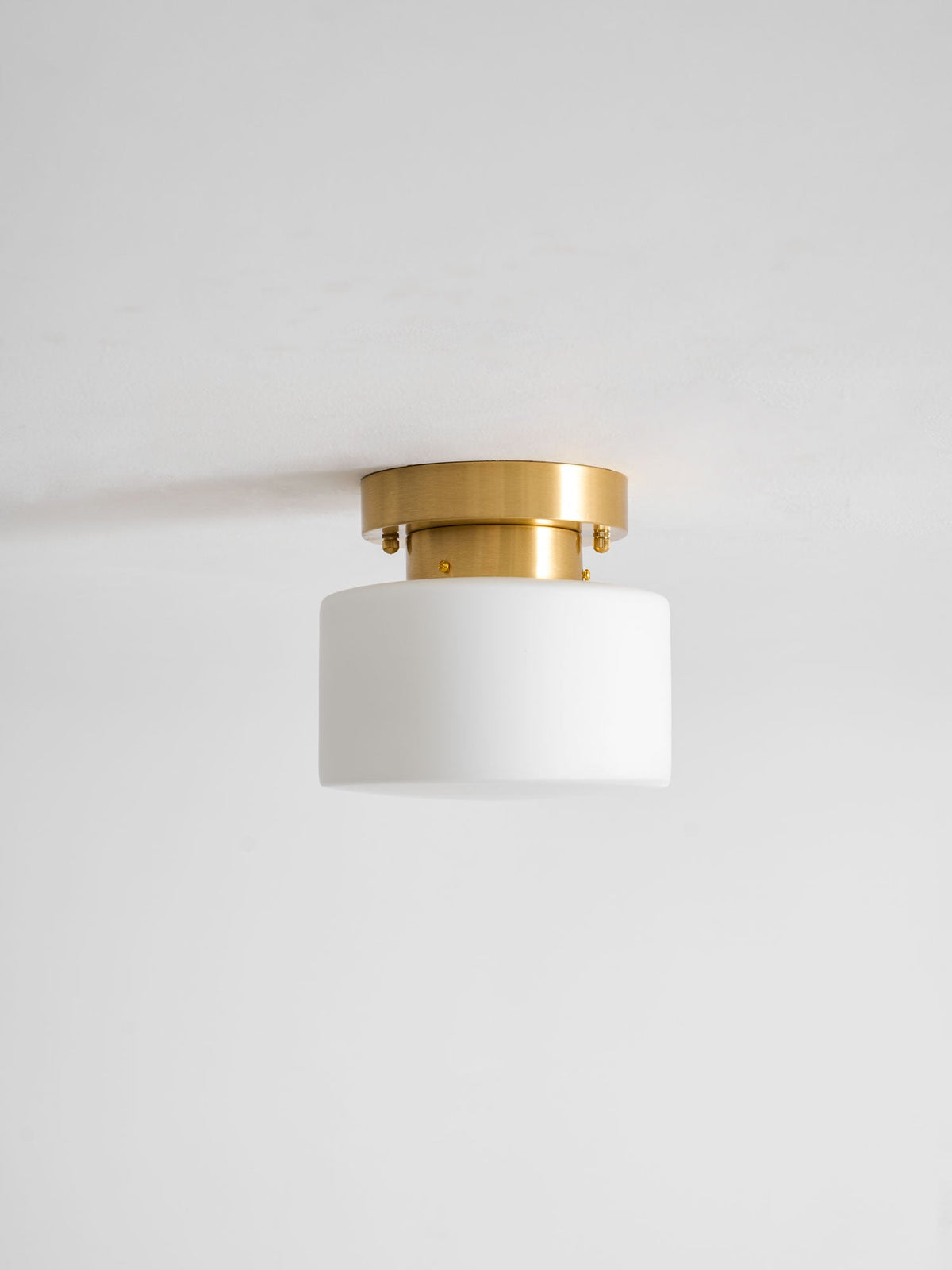 Unlit Clérachic flush-mount light with a white glass drum shade and brushed brass canopy against a pale grey ceiling.