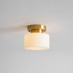 A lit Clérachic flush-mount light on a grey ceiling, the white glass drum shade glowing warm around the brass canopy.