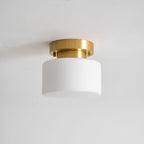 A Clérachic flush-mount light, unlit, with the white drum shade and round brass canopy seen from slightly below against grey plaster.