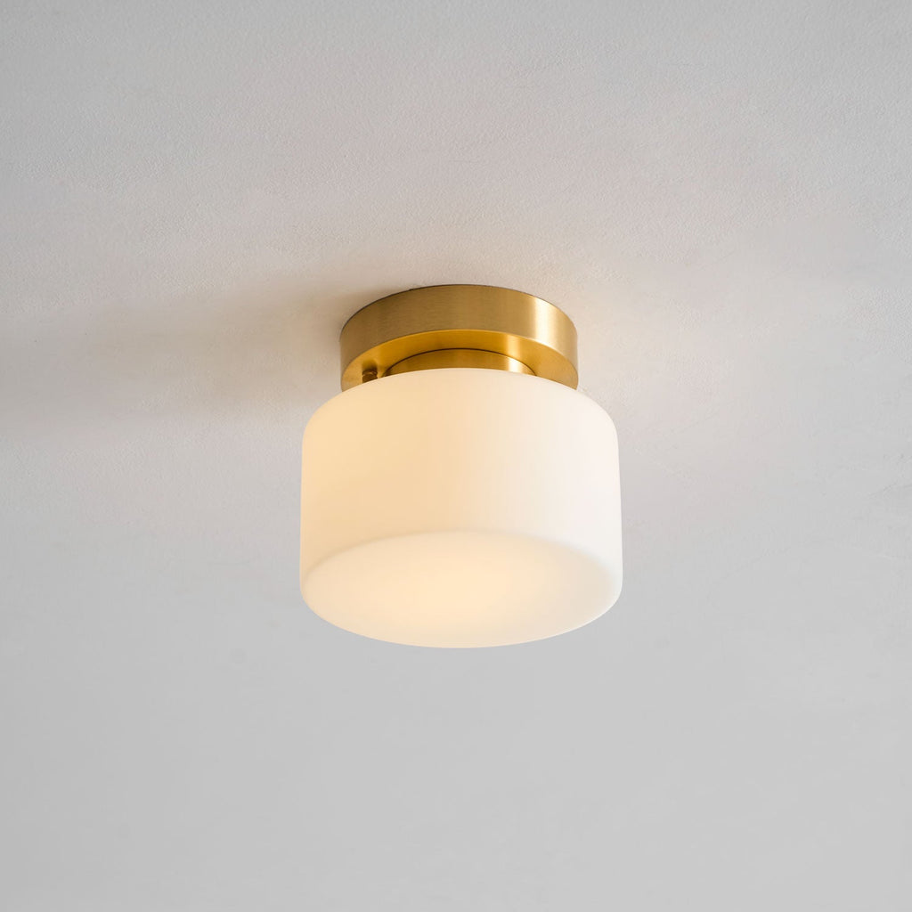 A Clérachic flush-mount light lit warmly, the frosted white shade glowing softly against a grey ceiling with a brass canopy above.