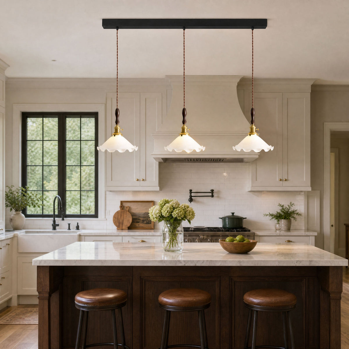 Three Clérachic pendant lights hang from a black bar over a marble-topped island with leather stools in a cream kitchen.