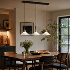 A row of three ruffled glass shades from this Clérachic pendant light hangs above a timber dining table with black cane chairs.