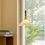 A Clérachic pendant light hangs in front of a blinded window with green leaves outside, above a stack of books.
