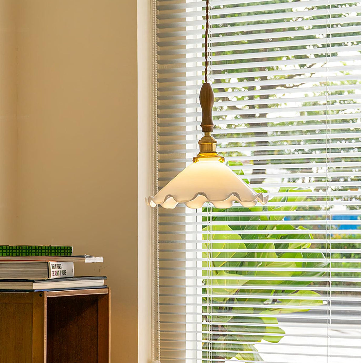 A Clérachic pendant light hangs in front of a blinded window with green leaves outside, above a stack of books.