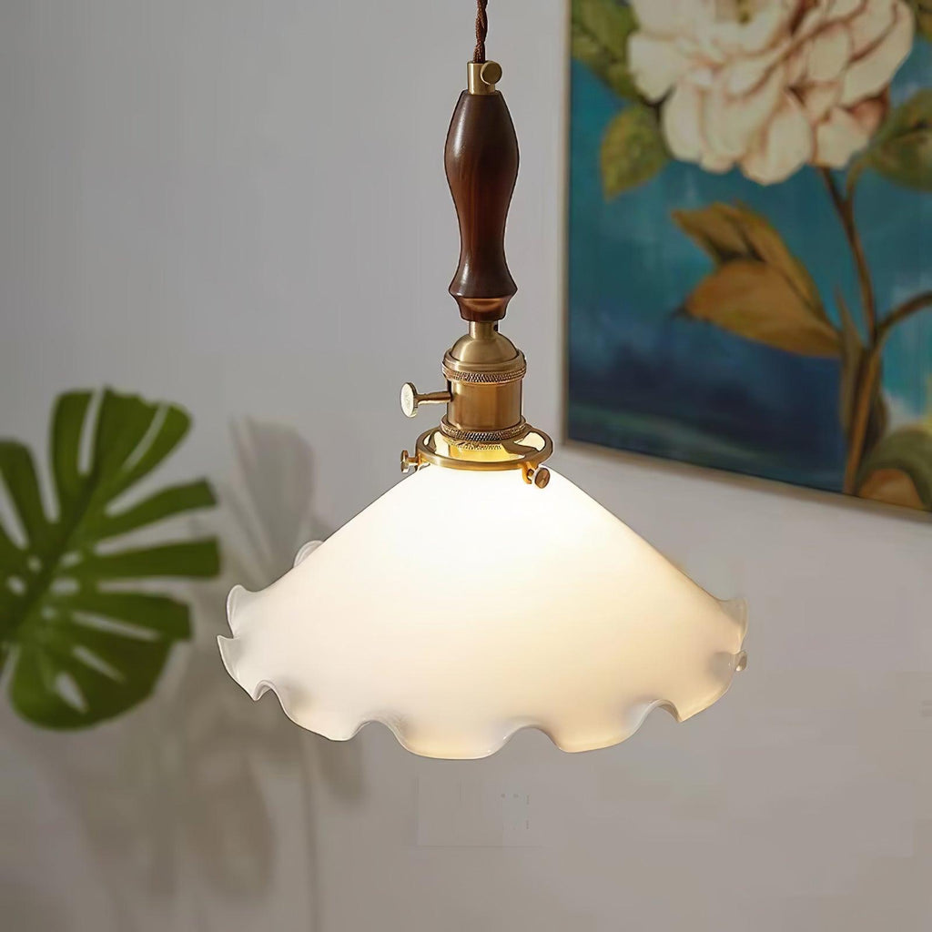 Close view of this Clérachic pendant light lit warmly, with a brass socket and switch above the frilled shade, a monstera leaf to the left.