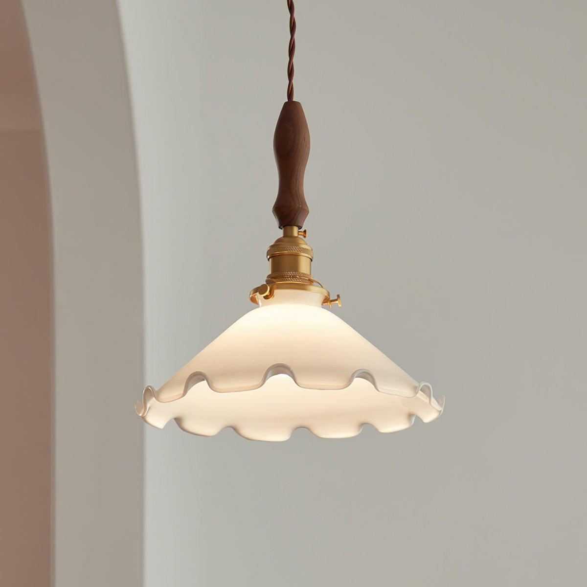 The frilled white shade of this Clérachic pendant light glows softly beside an arched wall, its walnut handle above brass fittings.