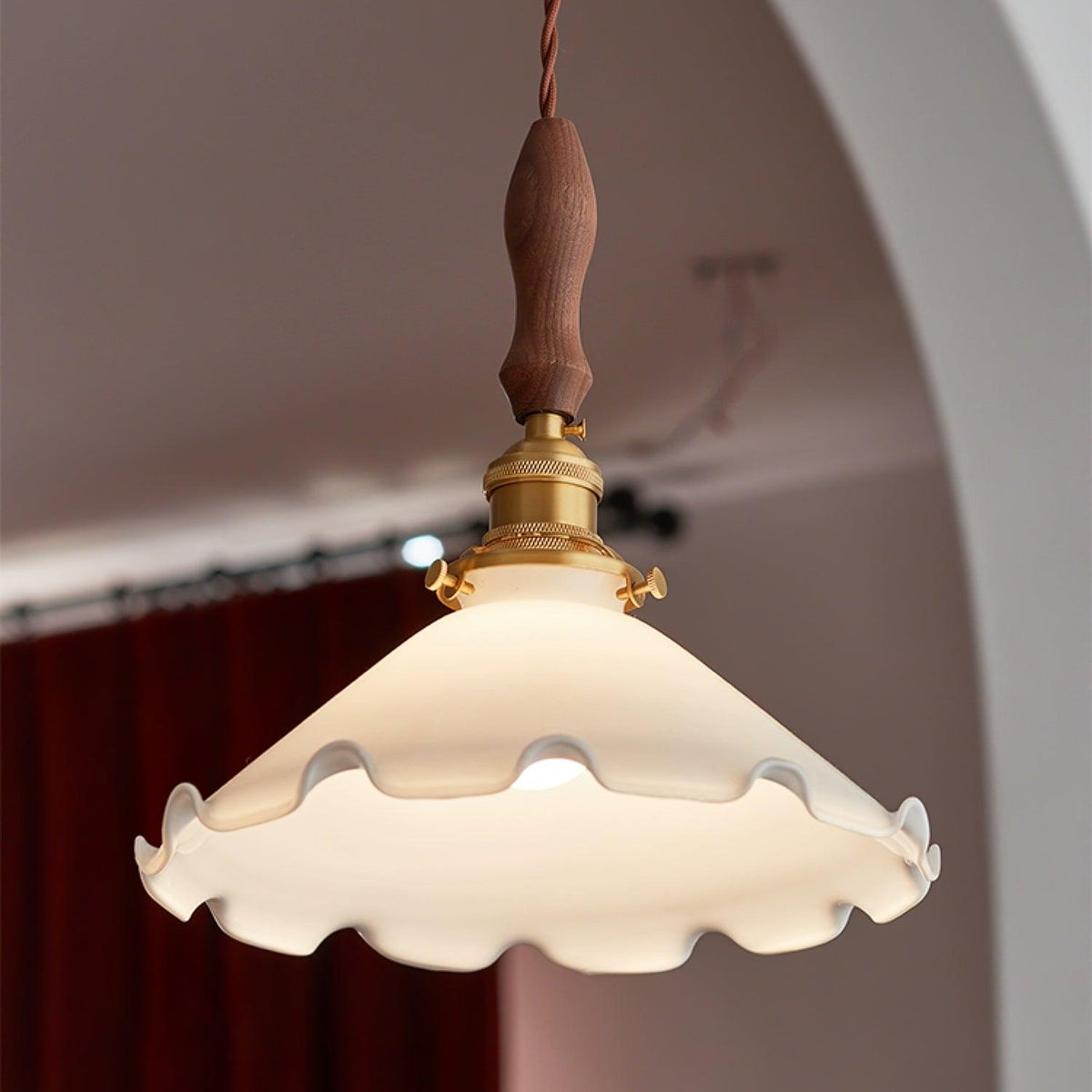 Looking up at this Clérachic pendant light, a wavy-edged white shade lit from within against an arched wall and a red curtain.