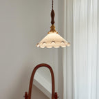 This Clérachic pendant light is lit against a pale wall, with sheer curtains to the right and a wooden mirror frame below.