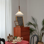 A small Clérachic pendant light hangs high above a red-clothed table set with a teapot, with a teak chest and palm behind.