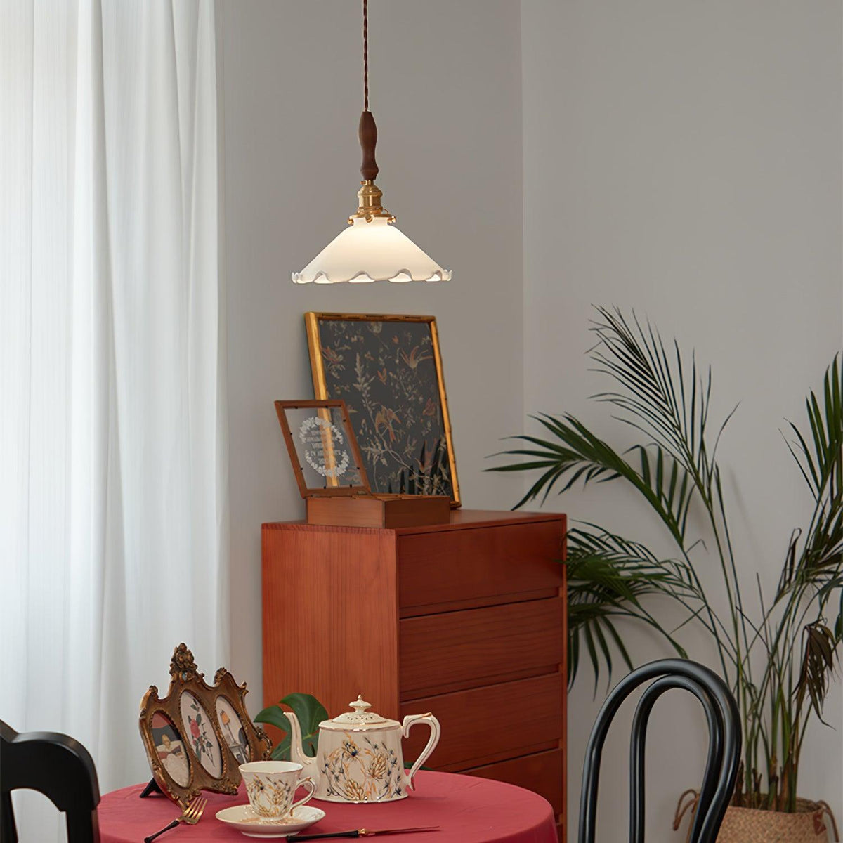 A small Clérachic pendant light hangs high above a red-clothed table set with a teapot, with a teak chest and palm behind.