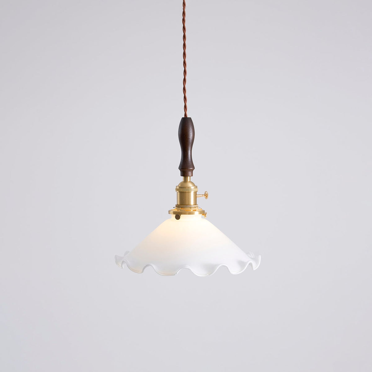 This Clérachic pendant light hangs against a pale grey wall, its white shade lit gently around the brass socket.