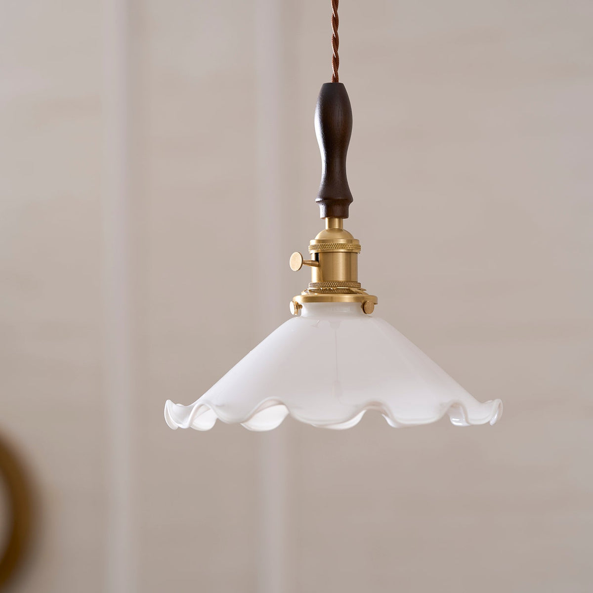 This Clérachic pendant light hangs against a beige wall, with a brass socket, turn switch and a ruffled white shade.